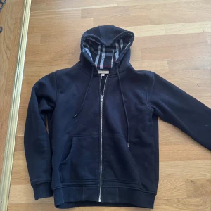 Svart hoodie från Burberry med dragkedja - Svart hoodie från Burberry med klassiskt rutigt foder i huvan och hel dragkedja framtill. Tröjan har snörning i huvan, känguruficka och ribbade muddar. Perfekt för dig som gillar stilren design med en touch av lyx.