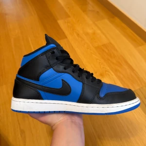 Nike Air Jordan 1 Mid Svart/Blå - Nike Air Jordan 1 Mid i svart och blått skinn med klassisk svart swoosh och blå yttersula. Skorna har snörning, rund tå och vit mellansula. Ikonisk Jordan Wings-logga på sidan och stilren design som sticker ut.