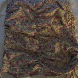 Blommig kortkjol med volang - Supersöt kortkjol med blommigt mönster i rosa, grön och gul. Kjolen har volangdetaljer och smockad struktur som ger en trendig look. Perfekt för dig som gillar färg och vill sticka ut lite extra.