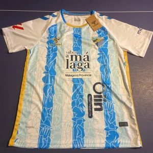 Fotbollströja  - Snygg Málaga CF fotbollströja i storlek M från Hummel i vitt och blått med unikt grafiskt mönster och gula detaljer på sidorna. Kortärmad modell i lätt och ventilerande funktionsmaterial. Klubblogga och sponsortryck på bröstet. Helt ny, prislapp sitter på!