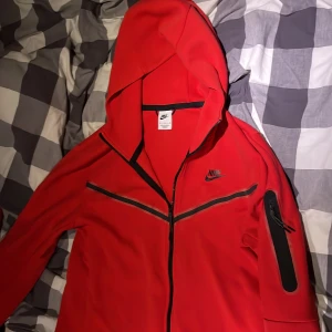 Röd Nike vindjacka med huva M - Nike vindjacka i rött med svarta detaljer och dragkedja. Jackan har huva, lång ärm och en snygg ficka med dragkedja på ena ärmen. Perfekt för dig som gillar sportig stil och vill sticka ut med färg.