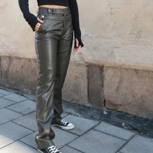 Mörkgröna skinnbyxor från A Dsgn - Snygga mörkgröna skinnbyxor från A Dsgn i rak modell. Byxorna har hög midja, bälteshällor och dragkedja framtill. Materialet är syntetiskt skinn med mjuk insida, perfekta för en edgy streetstil.