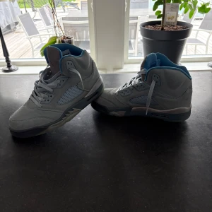 Nike Air Jordan 5 UNC blå sneakers - Nike Air Jordan 5 UNC i ljusblå mocka med meshpaneler och ikonisk Jumpman-logga på plösen och hälen. Skorna har högt skaft, snörning och detaljer i blått och grått. Perfekta för dig som vill sticka ut med en klassisk och sportig look. Låda följer med!