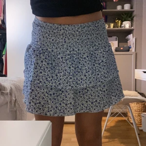 Blåvit blommig volangkjol - Supersöt kort kjol med blått blommönster på vit botten. Kjolen har smock i midjan och dubbla volanger som ger en flowy känsla. Perfekt för varma dagar och ger en fräsch sommarvibe. Materialet känns lätt och luftigt. Kjolen är i strl 152 från ginayoung men sitter som XS.