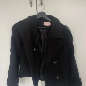 Svart skepparkavaj från Nelly - Stilren svart skepparkavaj från Nelly med dubbelknäppning och stora knappar. Jackan har en klassisk krage, axelklaffar och är tillverkad i ett ulligt material som ger en cool och tidlös vibe. Perfekt för dig som vill ha en snygg och enkel jacka till höst och vinter.