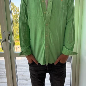 Grön skjorta från Polo Ralph Lauren - Snygg grön skjorta från Polo Ralph Lauren i custom fit. Skjortan har klassisk krage, knäppning framtill och lång ärm. Broderad orange logga på bröstet ger en clean och sportig vibe. Perfekt till jeans eller chinos för en fräsch look.