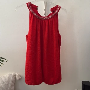 Röd pärlbroderad linnetopp från Gina - Snygg röd ärmlös linnetopp från Gina med pärlbroderier runt halsringningen. Toppen är gjord i 100% polyester och har en lätt rynkad struktur samt knäppning i nacken. Perfekt för dig som vill sticka ut med detaljer och färg.