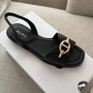 Svarta sandaler från ALDO med gulddetalj - Stilrena svarta sandaler från ALDO med bred rem och snygg guldfärgad metallspänne framtill. Platt sula och öppen tå, perfekt för varma dagar. Tillverkade i syntetmaterial med en elegant och modern look.