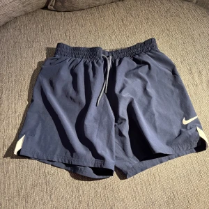 Blå Nike badshorts med snörning - Mörkblå badshorts från Nike med elastisk midja och snörning för perfekt passform. Klassisk vit Nike-logga på ena benet och vita detaljer vid bensluten.  Storlek XS