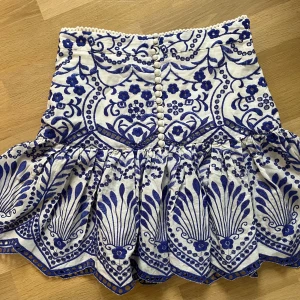 Vit och blå broderad kortkjol - Superfin kortkjol i vitt med blått broderat blommönster och dekorativa knappar framtill. Kjolen har volangig nederkant.