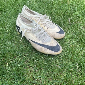Nike Mercurial fotbollsskor beige/svart - Säljer ett par Nike Mercurial fotbollsskor i beige med svarta detaljer och snörning. Skorna har en lätt ovandel i syntetmaterial och mönstrad sula med dobbar för bra grepp på planen. Perfekta för dig som vill ha snabba och stilrena skor på matchen.