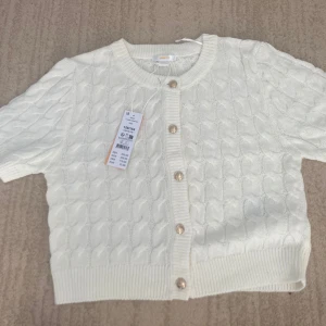 Vit kabelstickad topp från Gina Tricot - Superfin vit kabelstickad topp från Gina Tricot med korta ärmar och snygga guldfärgade knappar framtill. Toppen har rund halsringning och ribbade muddar vid ärmslut och nederkant. Perfekt för dig som gillar en klassisk men trendig look. 