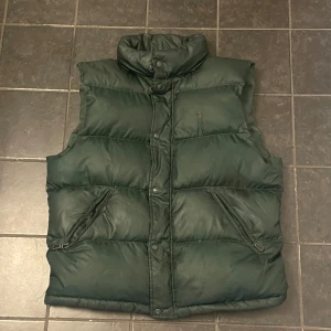 Mörkgrön dunväst från Polo Ralph Lauren - Dunväst i mörkgrönt från Polo Ralph Lauren med broderad logga på bröstet. Västen har två snedställda fickor med dragkedja och knäppning framtill. Klassisk quiltad design och huva som man kan veckla ut ur kragen för extra funktion.