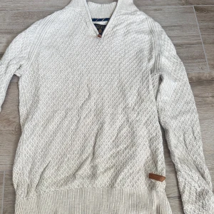 Vit stickad tröja med half zip Bondelid - Säljer en vit stickad tröja från Bondelid i storlek S. Tröjan har ett snyggt strukturmönster, ribbade muddar och krage samt en half zip-dragkedja med läderdetalj. Perfekt för dig som gillar klassisk och stilren look.