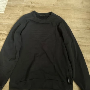 Svart sweatshirt från Emporio Armani - Svart sweatshirt från Emporio Armani med det klassiska logotypen ton-i-ton på bröstet. Tröjan har rund halsringning och långa ärmar. Tillverkad i mjuk bomull som är skön att bära. Perfekt för dig som gillar stilrena och exklusiva plagg.