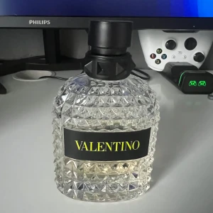 Valentino parfymflaska med nitar - Säljer en lyxig parfym från Valentino i en transparent glasflaska med coolt nitat mönster och svart kork med logga. Flaskan har en tydlig etikett med gult tryck. Perfekt för dig som gillar exklusiva dofter och snygg design på hyllan.100 ml flaska men de finns ba cirka 30 ml kvar 