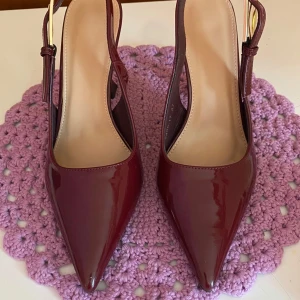 Vinröda lackpumps med spetsig tå - Snygga vinröda pumps i lack med spetsig tå och hög klack. Skorna har öppen häl med justerbart spänne i guld och en elegant, smal siluett. Perfekta för dig som vill sticka ut med en trendig och stilren look.