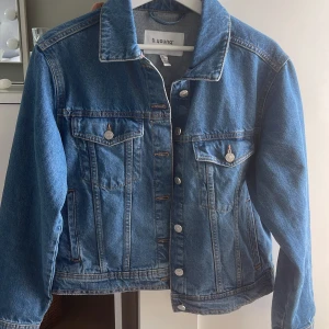 Blå jeansjacka från b.young - Snygg blå jeansjacka från b.young med klassisk passform, raka linjer och silverfärgade knappar. Jackan har två bröstfickor med lock och knappar samt långa ärmar. Perfekt att slänga över en hoodie eller t-shirt för en chill look. Den är helt ny och har aldrig används. Säljer då jag fick en annan. Normalpris 590kr. 