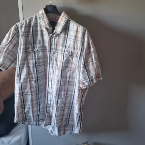 Rutig kortärmad skjorta från Blend XXL - Snygg rutig skjorta från Blend i storlek XXL. Skjortan är vit med inslag av orange, blått och svart i rutmönstret. Den har korta ärmar, klassisk krage, två bröstfickor med knappar och stängs med knappar framtill. Perfekt för en chill och avslappnad stil.