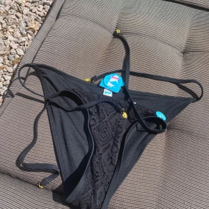 Svart bikini underdel med spets och gulddetaljer str M - Supersnygg svart bikini med spetsdetaljer och tunna snören med små guldfärgade löv. Bikinin har knyt i sidorna och en elegant, feminin look. Perfekt för stranden eller poolen när du vill sticka ut lite extra.