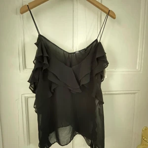 Svart volangtop från Zara - Svart, tunn och flowy linnetopp från Zara med smala axelband och flera lager volanger framtill och längs sidorna. Toppen är lätt transparent och har en luftig känsla, perfekt för att skapa en cool och trendig look.
