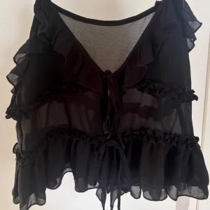 Svart linne med volanger och knyt - Supersöt svart linne med volanger längs v-ringningen och över hela toppen. Knytning framtill ger en extra detalj och den är luftig och flowy. Perfekt för dig som gillar romantisk och trendig stil.