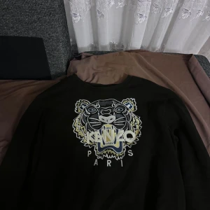 Svart Kenzo sweatshirt med tigerbrodyr - Svart sweatshirt från Kenzo med ikonisk tigerbrodyr och texten 'KENZO PARIS' framtill. Tröjan har rund halsringning och långa ärmar. Brodyren har detaljer i vitt, blått och gult som ger en snygg kontrast mot den svarta basen.