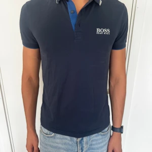 Marinblå pikétröja från Hugo Boss - Snygg marinblå pikétröja från Hugo Boss med vit logga på bröstet. Tröjan har klassisk krage med blå insida och vita detaljer på kragen. Kortärmad och tillverkad i mjuk bomull, perfekt för en stilren look.