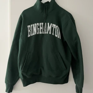 Mörkgrön Binghamton half zip sweatshirt från champion  - Mörkgrön sweatshirt med vit BINGHAMTON-text över bröstet från champion . Tröjan har half zip i halsen, två fickor framtill och en relaxed passform. Perfekt för chill dagar eller när du vill ha en sportig vibe.