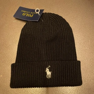 Svart ribbad mössa från Polo Ralph Lauren - Snygg svart ribbad mössa från Polo Ralph Lauren med broderad logga framtill. Perfekt för kyliga dagar och en stilren look.