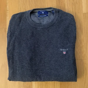 Mörkblå stickad tröja från GANT - Snygg mörkblå stickad tröja från GANT med rund halsringning och diskret logga broderad på bröstet. Tröjan är gjord i bomull och har en klassisk, stilren look som passar perfekt till jeans eller chinos.