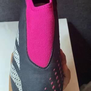 Adidas Predator fotbollsskor rosa/svart - Adidas Predator fotbollsskor i svart och stark rosa med silvriga detaljer. Skorna har en strumpliknande ovandel i syntet och textil, samt mönstrad yta för extra grepp på planen. Ikoniska tre ränder på sidan och låg profil för snabbhet.
