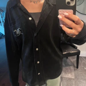 Svart Juicy Couture skjorta i velour - Svart långärmad skjorta från Juicy Couture i mjuk velour med glittrande stenar på bröstfickan och stor Juicy Couture-logga i strass på ryggen. Skjortan har klassisk krage och knäppning framtill med silvriga knappar. Perfekt för dig som gillar statement-plagg.