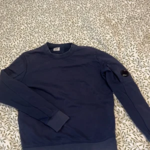 Blå sweatshirt från C.P. Company - Säljer en mörkblå sweatshirt från C.P. Company i bomull. Tröjan har rund hals, långa ärmar och den ikoniska linsen på ena ärmen. Perfekt för dig som gillar stilrena och snygga plagg med en twist.