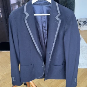 Mörkblå kavaj Lanvin x H&M 2010 - Snygg svart kavaj från Lanvin x H&M kollektionen 2010. Kavajen har grå kontrastkant längs slaget, tre knappar framtill och två fickor. Fodrad insida för extra komfort och stilren look. Perfekt för dig som vill sticka ut med en unik designerpiece.