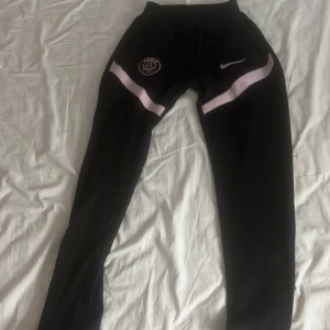 Svarta PSG träningsbyxor Nike - Svarta träningsbyxor från Nike med PSG-logga på låret och vita detaljer över låren. Byxorna har smal passform och är perfekta för träning eller sport. Elastisk midja och mjukt, syntetiskt material som andas.
