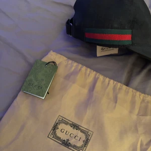 Svart keps från Gucci med röd-gröna detaljer - Exklusiv svart keps från Gucci med klassiska röd-gröna ränder på sidan. Justerbar rem baktill för perfekt passform. Kepsen är tillverkad i bomull och har diskret GG-mönster i tyget. Kommer med originalpåse och tagg.