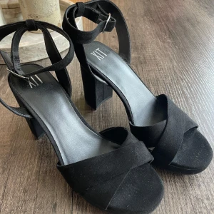 Svarta klackade sandaler från XIT - Använda 1 gång! Svarta sandaler från XIT med bred klack och öppen tå. Remmar i mockaimitation som korsas framtill och justerbar ankelrem med silverfärgat spänne. Perfekta för dig som vill ha en stilren och trendig look. Stl 38 