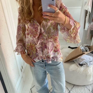 Blommig och trendig volangblus  - Säljer en superfin blus med blommigt mönster i rosa, lila och grönt från Zara. Blusen har volangdetaljer längs v-ringningen och ärmarna, samt luftiga trekvartsärmar. Perfekt för dig som gillar romantisk och bohemisk stil.