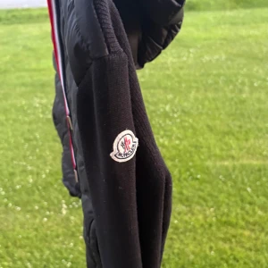 Svart Moncler pufferjacka med huva - Svart pufferjacka från Moncler med quiltad framsida, stickade ärmar och huva. Jackan har dragkedja framtill och coola röda, vita och blå snören. Moncler-logga på ärmen ger extra premiumkänsla. Perfekt för dig som vill ha en stilren och sportig look.