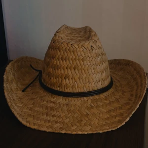 Houston II Straw Cowboyhatt S/M - Still ren cowboyhatt i flätad halm med bred brätte och klassisk form. Hatten är ljusbrun (tan) och har ett mörkbrunt band runt kronan som knyts i ena sidan. Perfekt för dig som vill sticka ut med westernstil.