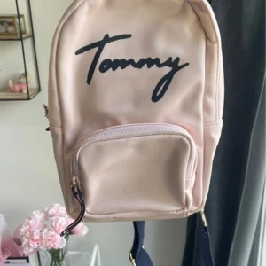 Rosa ryggsäck Tommy Hilfiger - Snygg ljusrosa ryggsäck från Tommy Hilfiger med mörkblå logga framtill. Väskan har ett stort huvudfack, en mindre ficka med dragkedja framtill och marinblått innerfoder med röda detaljer. Justerbara axelband och guldfärgade metalldetaljer.