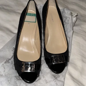 Svarta wedgepumps från Calvin Klein - Snygga svarta pumps från Calvin Klein med kilklack och öppen tå. Skorna har ett diskret mönster i tyget och en lackad detalj framtill med CK-logga i metall. Bekväm innersula i beige och klassisk rundad form. Perfekta för dig som gillar stilrena detaljer.