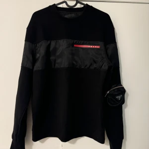 Svart Prada sweatshirt med ficka - Svart sweatshirt från Prada med cool nylonpanel över bröstet och röd logodetalj. Unik ficka med dragkedja och metallogo på ena ärmen. Långärmad och i en mix av bomull och nylon för en modern vibe. Endast testad. Storlek S men passar även M.