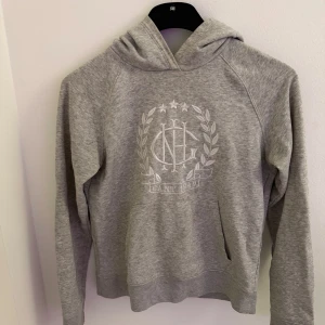 Grå hoodie från Gant med tryck - Snygg grå hoodie från Gant med klassiskt emblemtryck framtill. Tröjan har huva, långa ärmar och en känguruficka. Perfekt för en chill och sportig look. Mjuk bomullsblandning som känns skön mot huden.
