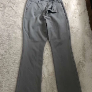Grå kostymbyxor med bootcut - Snygga grå kostymbyxor med bootcut-snitt och klassiska fickor. Byxorna har hällor för bälte och stängs med dragkedja och knapp. Perfekta för en clean och stilren look. Passar 38-40