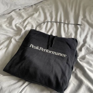 Svart Peak Performance hoodie - Svart hoodie från Peak Performance med vit logga tryckt över bröstet. Klassisk modell med huva och snörning, tillverkad i mjuk bomull. Perfekt för en chill och avslappnad stil.