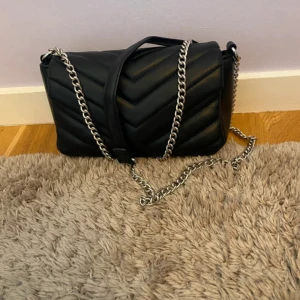 Svart quiltad axelväska med kedja - Snygg svart axelväska med quiltad design och silvrig kedjerem. Väskan har ett lock med magnetknäppning och ett rymligt huvudfack. Perfekt storlek för mobil, plånbok och andra essentials. Stilren och trendig accessoar som lyfter varje outfit.