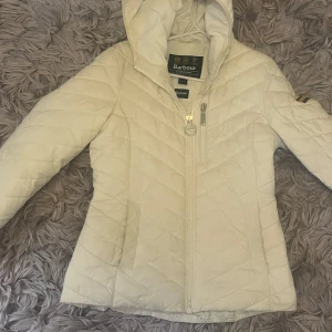 Snygg vit jacka från barbour - Snygg vit pufferjacka från Barbour International med huva och quiltad design. Jackan har dragkedja framtill, en bröstficka med dragkedja och Barbour-logga på ärmen. Perfekt för kyliga dagar när du vill vara både varm och stilren.