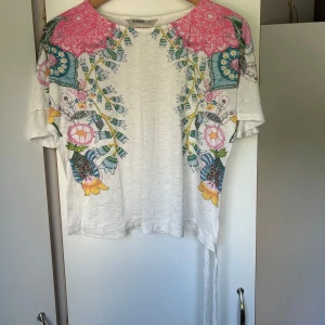 Vit blommig topp från Desigual - Superfin vit topp från Desigual med färgglada blommiga mönster i rosa, blått, gult och grönt. Toppen har korta ärmar och rund halsringning. Perfekt för dig som gillar unika och färgstarka plagg!